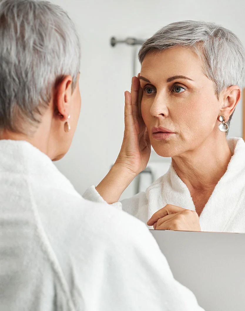 A woman with short gray hair in a white robe examines her reflection in a mirror, touching her face thoughtfully. - FaceLift in West Chester Pike Newton Square, PA