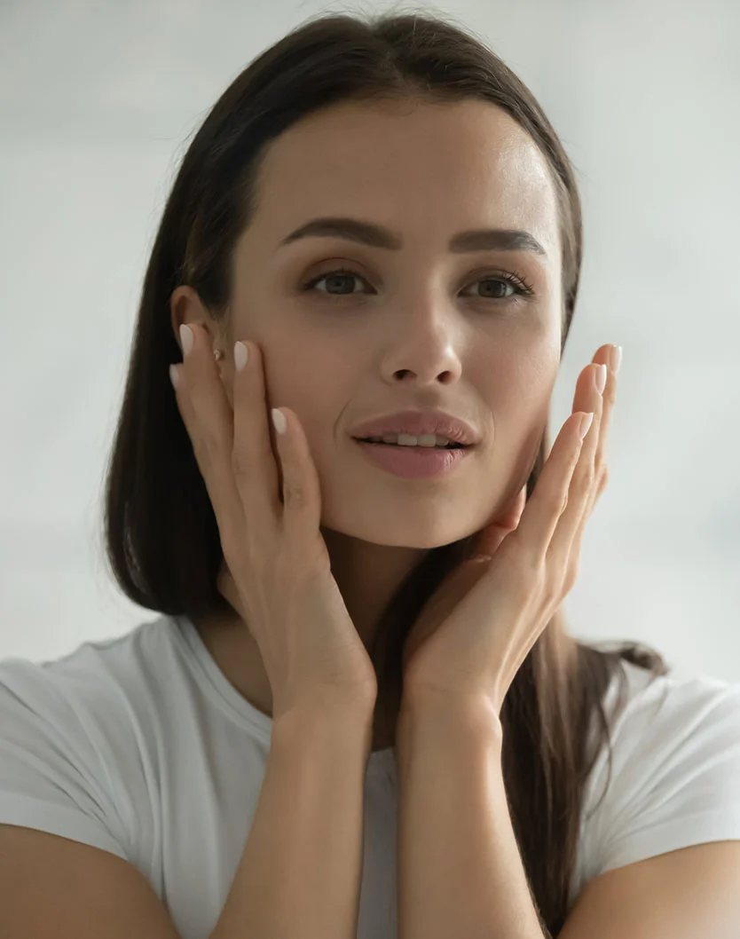 A young woman gently touches her face, with a calm expression, wearing a white shirt against a soft blurred background. - HydraFacial in West Chester Pike Newton Square, PA