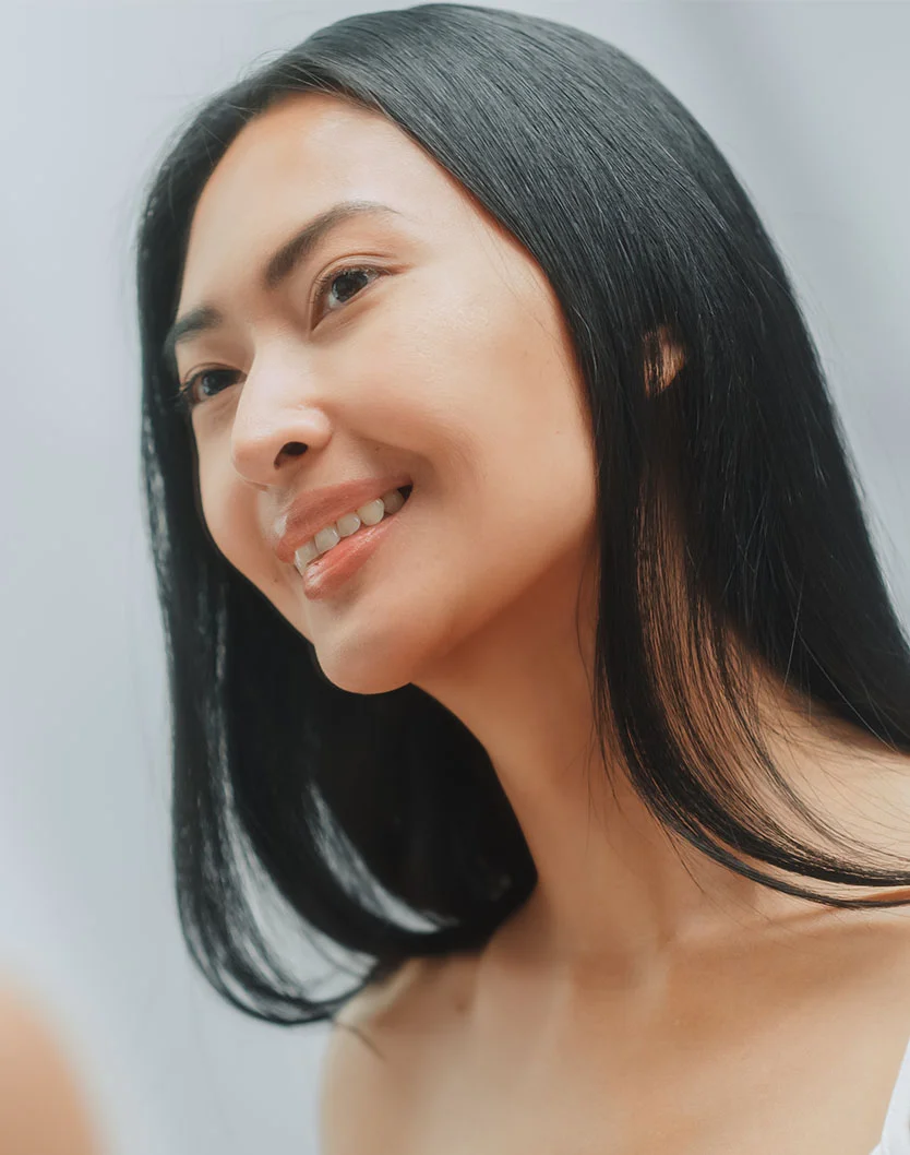 A smiling woman with long, straight black hair looks to the side, her soft features illuminated by natural light. - Lower Eyelid Surgery in West Chester Pike Newton Square, PA