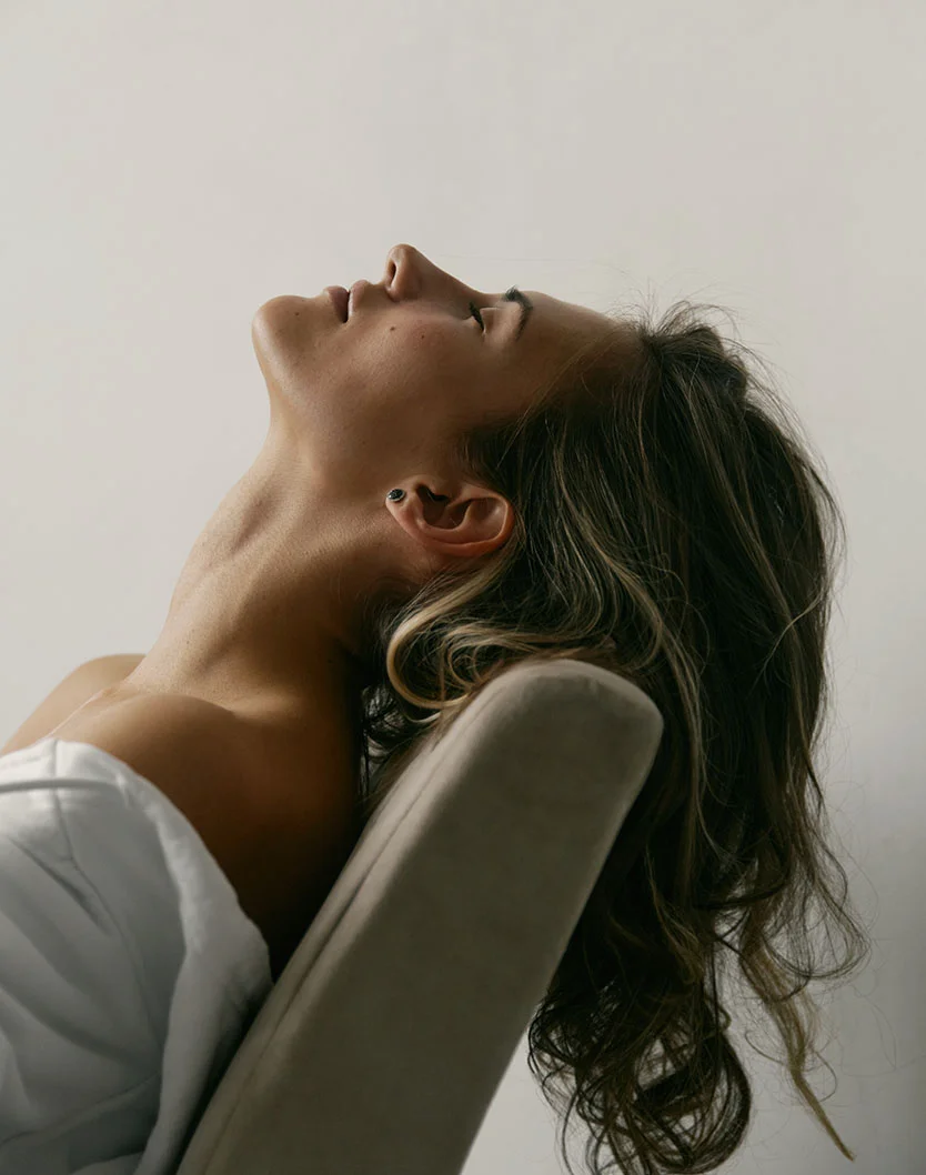 A woman with wavy hair leans back in a chair, eyes closed, with a serene expression against a plain background. - Neck and Chin Liposuction in West Chester Pike Newton Square, PA