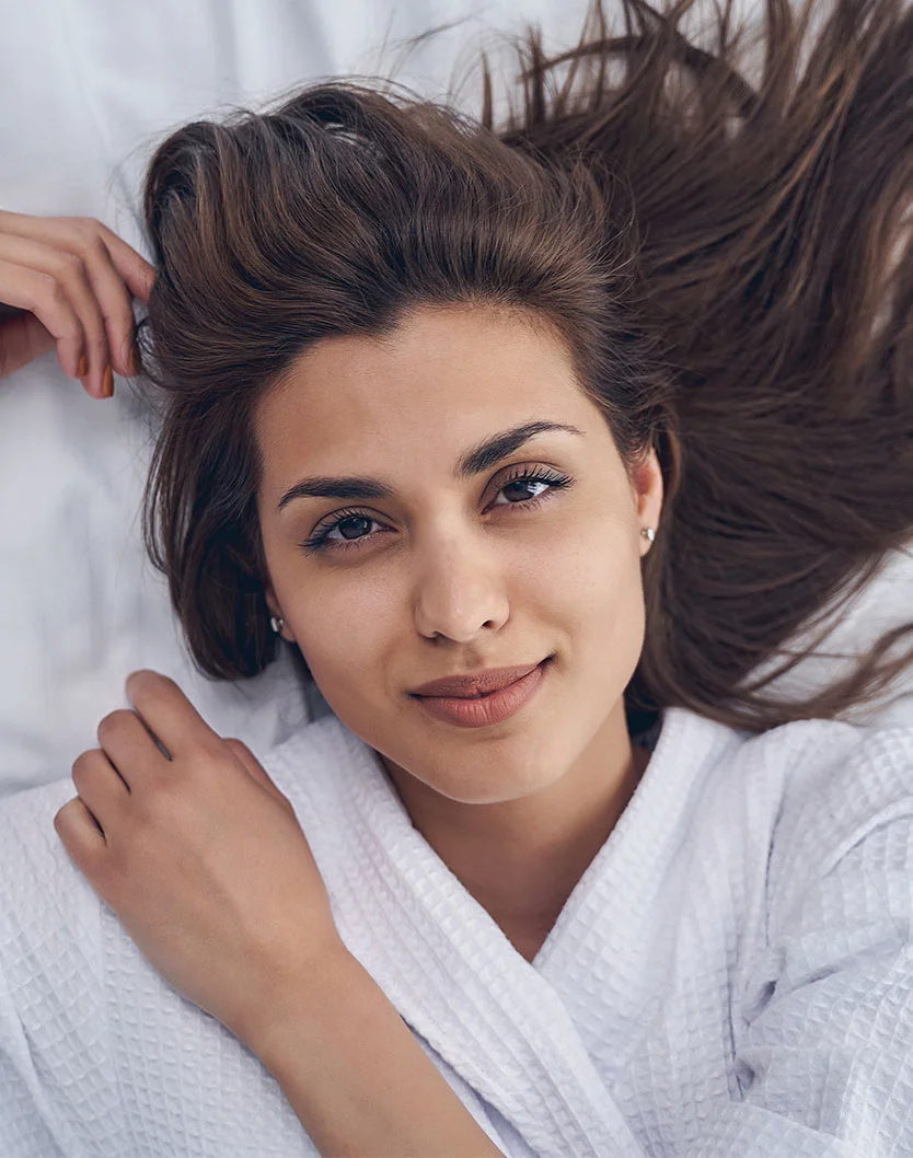 A young woman with long brown hair lies on a white bed, wearing a white robe, smiling softly at the camera. - Sculptra in West Chester Pike Newton Square, PA