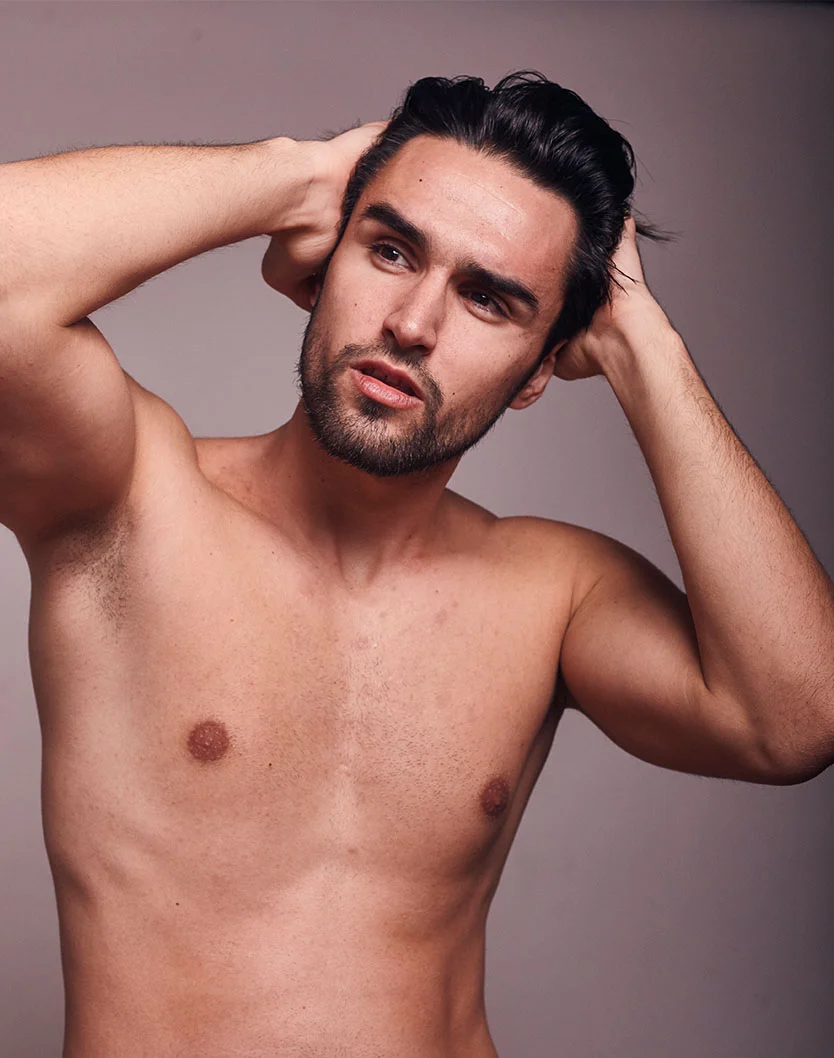 A young man with dark hair and a beard poses shirtless, holding his hair back, against a neutral background. - Top Surgery in West Chester Pike Newton Square, PA