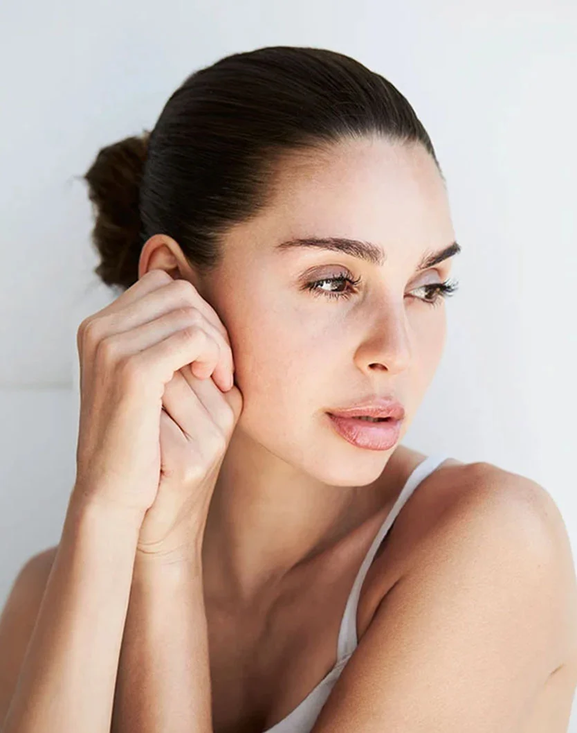 A woman with long hair in a bun gazes thoughtfully to the side, her hand resting on her cheek against a light background. - Upper Eyelid Surgery in West Chester Pike Newton Square, PA
